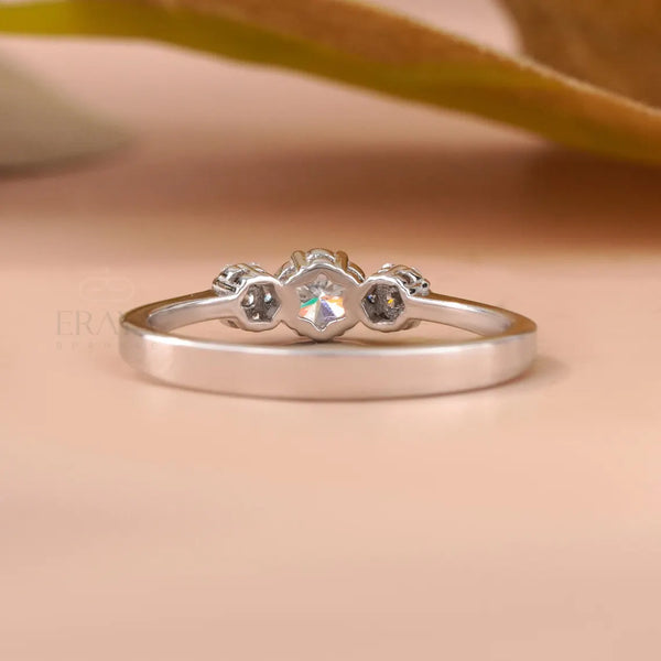 A silver ring with a central clear gemstone and two small dark gemstones on either side, resting on a soft beige surface with a blurred leaf in the background.
