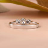 A silver ring with a central clear gemstone and two small dark gemstones on either side, resting on a soft beige surface with a blurred leaf in the background.
