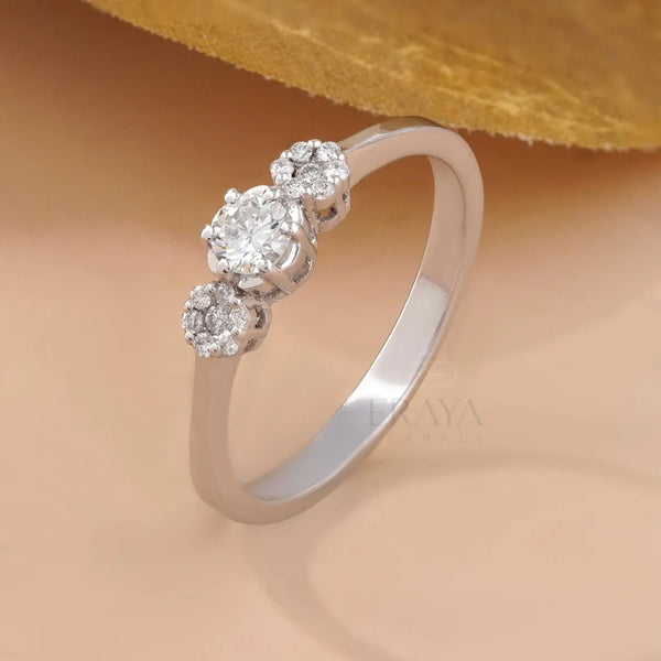 A silver three-stone diamond ring with a polished band, featuring a central round diamond flanked by two smaller round diamonds, set against a warm beige background with a gold leaf at the top.