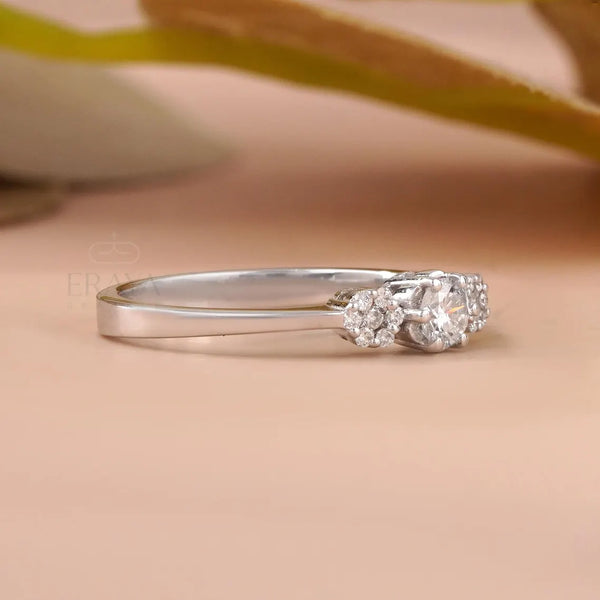 A silver three-stone diamond ring with a polished band and a cluster of three clear gemstones on the right side, resting on a soft beige surface with a blurred background.