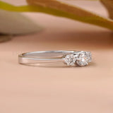 A silver three-stone diamond ring with a polished band and a cluster of three clear gemstones on the right side, resting on a soft beige surface with a blurred background.