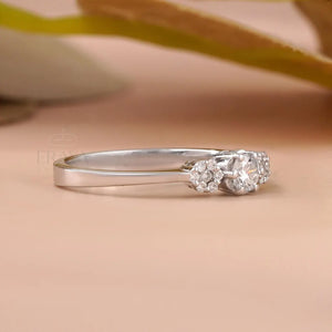 A silver three-stone diamond ring with a polished band and a cluster of three clear gemstones on the right side, resting on a soft beige surface with a blurred background.
