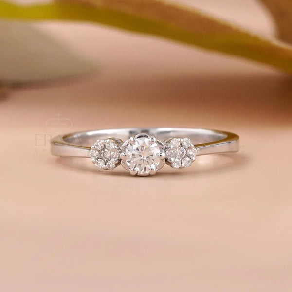 A silver three-stone diamond ring with a central round diamond and two smaller diamonds on either side, set against a soft peach background with a blurred olive branch.
