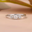 A silver three-stone diamond ring with a central round diamond and two smaller diamonds on either side, set against a soft peach background with a blurred olive branch.