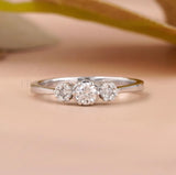 A silver three-stone diamond ring with a central round diamond and two smaller diamonds on either side, set against a soft peach background with a blurred olive branch.