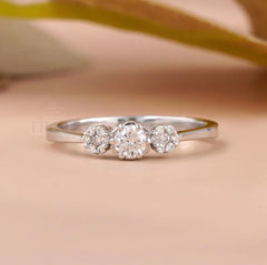A silver three-stone diamond ring with a central round diamond and two smaller diamonds on either side, set against a soft peach background with a blurred olive branch.