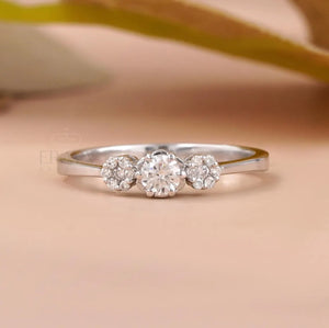A silver three-stone diamond ring with a central round diamond and two smaller diamonds on either side, set against a soft peach background with a blurred olive branch.