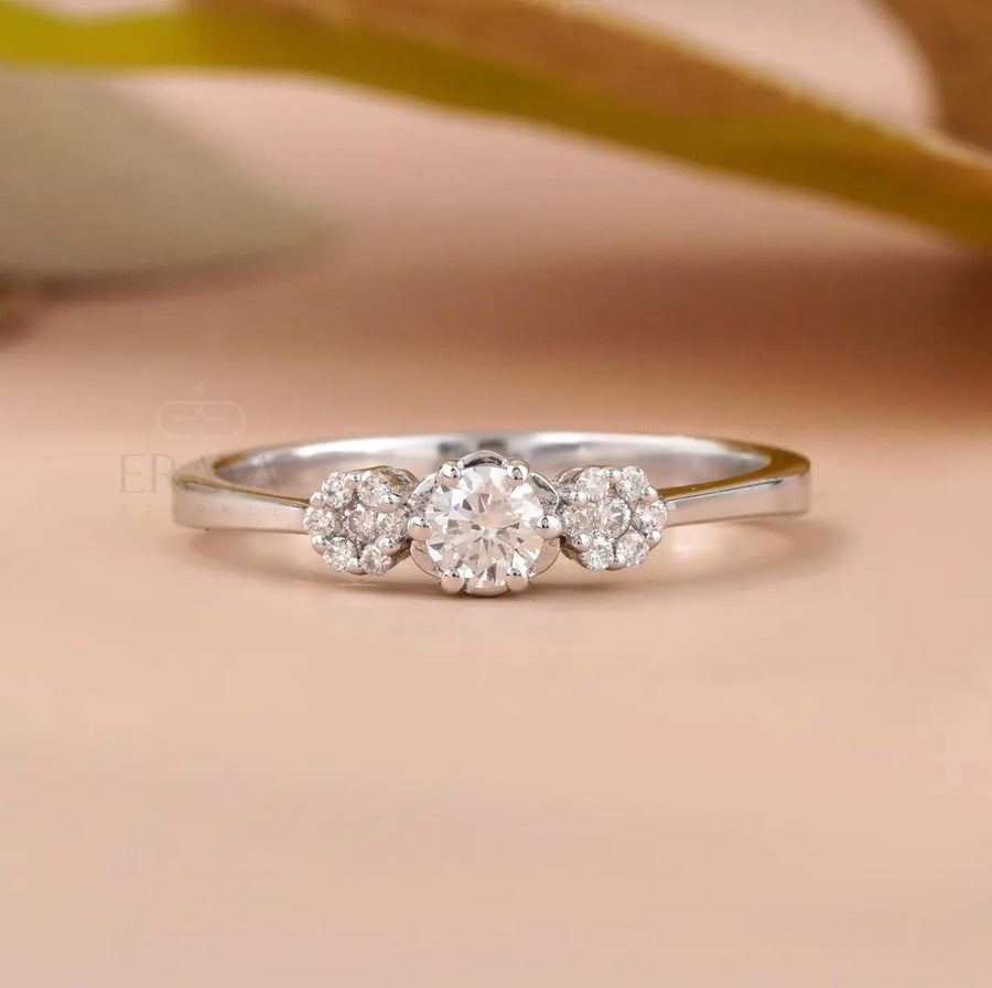 A silver three-stone diamond ring with a central round diamond and two smaller diamonds on either side, set against a soft peach background with a blurred olive branch.
