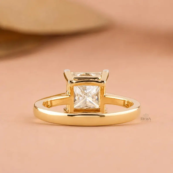 A gold ring with a princess-cut clear gemstone in a four-prong setting, displayed on a soft peach background.