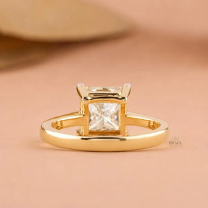 A gold ring with a princess-cut clear gemstone in a four-prong setting, displayed on a soft peach background.