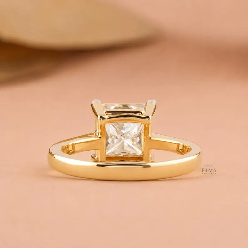 A gold ring with a princess-cut clear gemstone in a four-prong setting, displayed on a soft peach background.