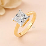 A gold ring featuring a large clear princess-cut diamond solitaire, set against a soft peach background with a blurred cream flower in the corner. The brand name 'ERAYA' is visible as a watermark on the ring's band.