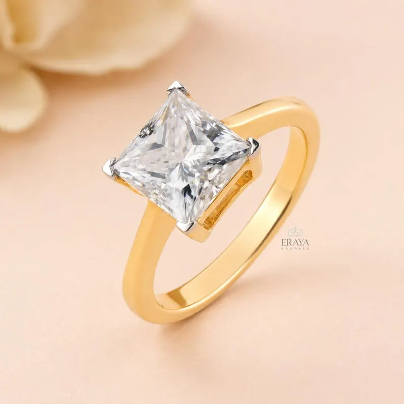 A gold ring featuring a large clear princess-cut diamond solitaire, set against a soft peach background with a blurred cream flower in the corner. The brand name 'ERAYA' is visible as a watermark on the ring's band.